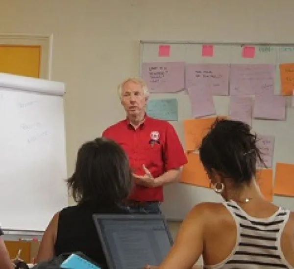 White man with white hair facing a group of young women sitting in chairs facing him, with a white paper on a tripod