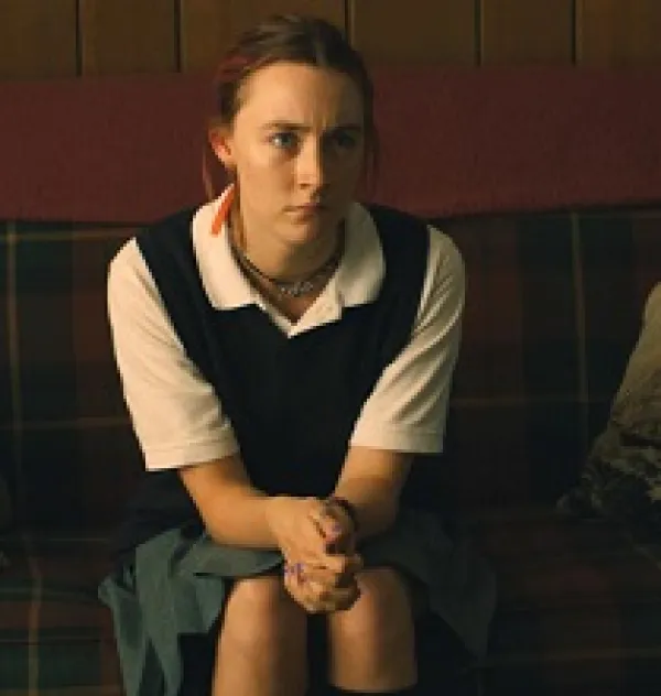 Young white woman in a white blouse, vest and hair pulled back sitting on a couch with hands clasped looking irritated
