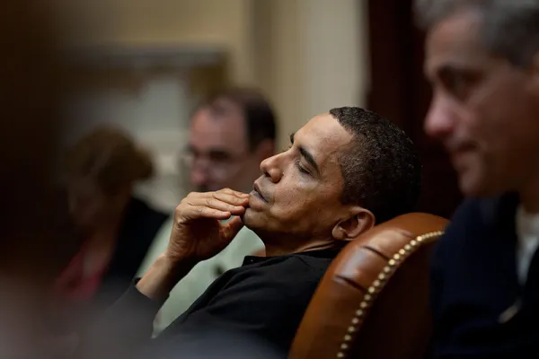 Obama leaning back in leather chair with hand on mouth looking pensive