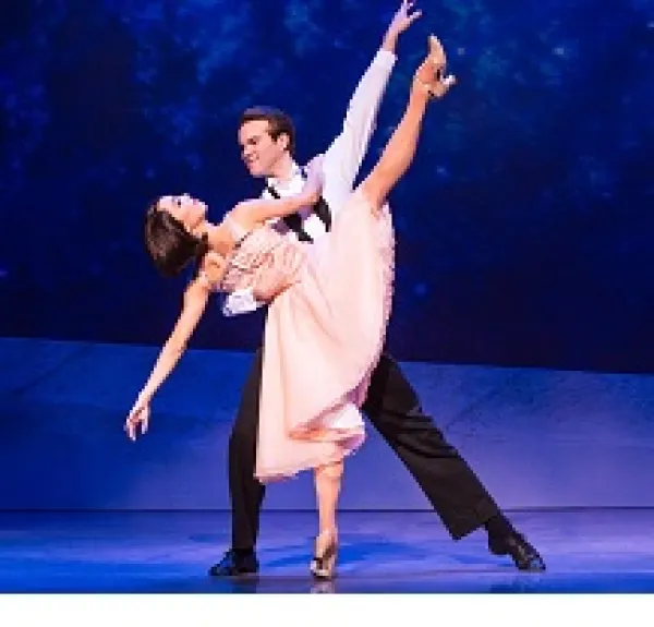 White man with dark hair in white shirt and black pants dancing with a white woman with short dark hair in a pick dress, he is leaning over as he holds her as she is kicking her leg high up in the air 