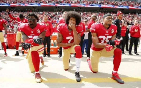 African American football players with red jerseys and tan pants kneeling on one knee in a stadium full of people