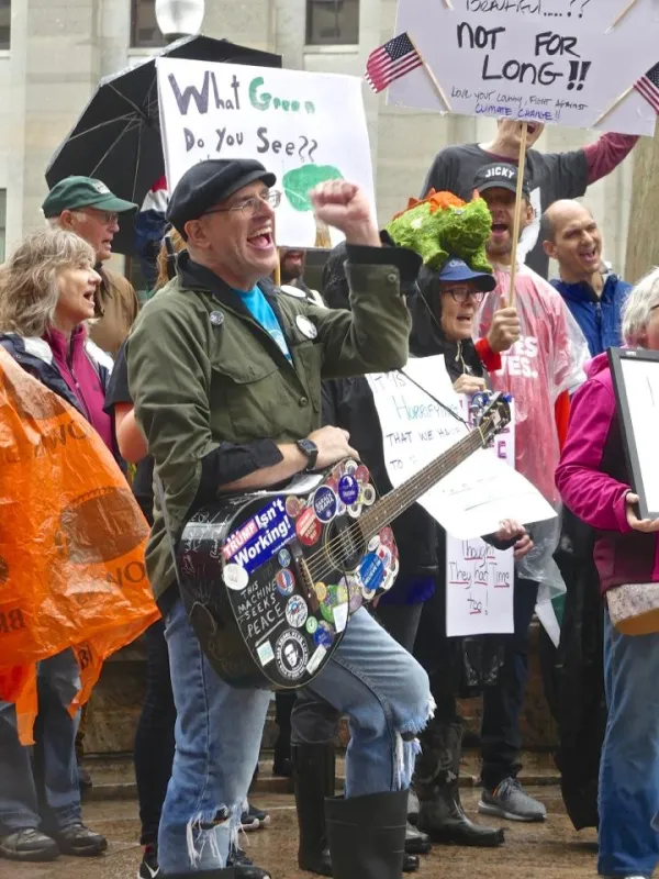 Guy with cap and guitar covered with stickers with his fist in the air in front of a crowd of people