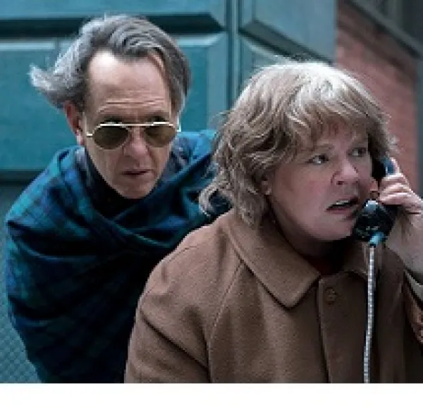 A man with sunglasses and hair blowing in the wind hovering behind a woman looking upset talking into a pay phone