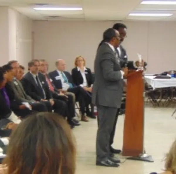 Lots of men in suits sitting in a row on chairs facing right and two black men in suits standing at a podium