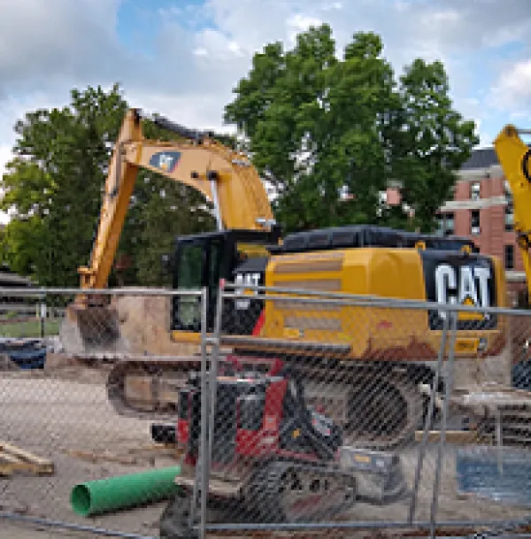 Big yellow machine truck with big scoop on a long neck that says CAT on the side behind a wire fence with other machinery and trees in the background