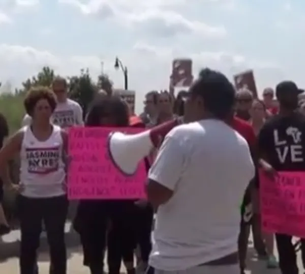 Man with brown hair and white shirt with back to camera holding a bullhorn surrounded by protestors