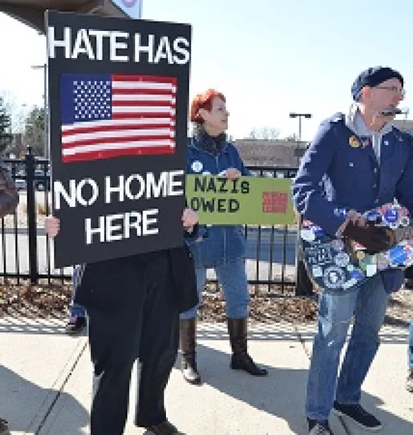 Someone holding a sign with a US flag on it on black background saying Hate has no home here, a guy playing a guitar that's covered with stickers with political sayings and a woman with red hair behind in the middle with a sign against Nazis