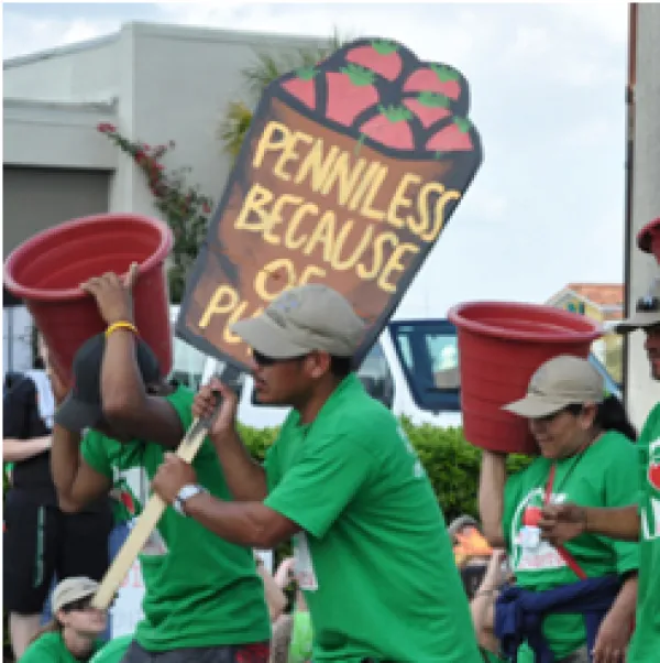 Dark-skinned men and women in green shirts at a demonstration marching to the left one holding a sign saying Penniless because of and two others holding big rust colored buckets