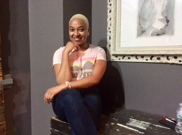 Young Black woman smiling and posing with her hand to her chin next to a painting