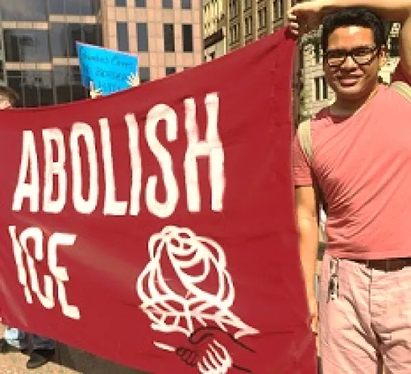 Young dark haired man with glasses holding a large red sign that says Abolish ICE with a DSA symbol of fist and rose