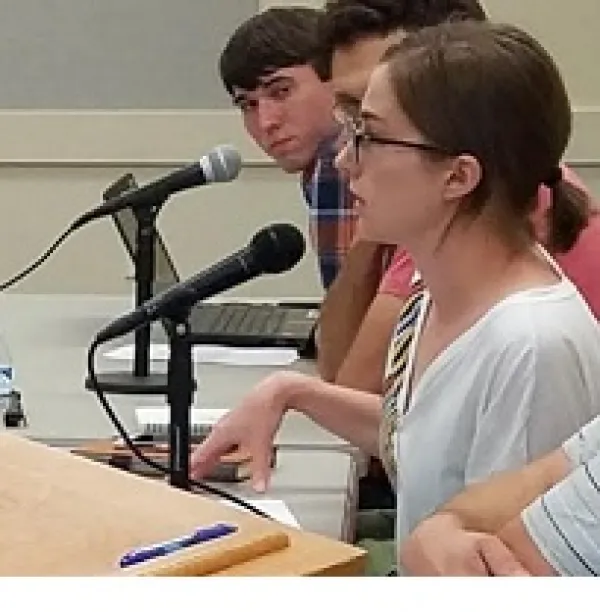 Young white woman with glasses and brown hair tied back speaking and gesturing at a podium with two guys at the table behind her 