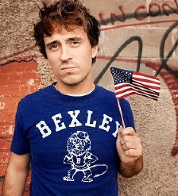 Young brown haired man wearing a blue t-shirt saying Bexley and waving a tiny flag in front of some kind of wall with words on it